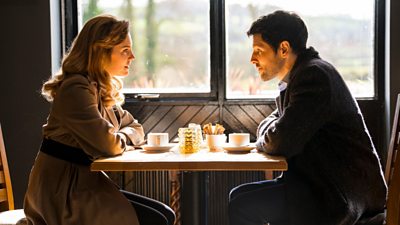 Cathy (Annabel Scholey) and Michael (Colin Morgan) sitting opposite one another at a table with cups and saucers in front of them 