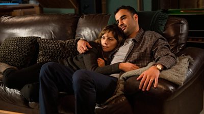 Raymie (Waj Ali) and Cathy (Annabel Scholey) cuddled up on a sofa looking at something offscreen
