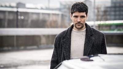 Michael (Colin Morgan) looking into the camera. he's wearing a t-shirt and winter coat. blurred city with snow in the background