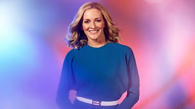 Gabby Logan smiling. She's wearing a blue long sleeved dress with a white belt. Background of blurred pastel pinks and purples