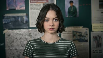 Pip Fitz-Amobi (Emma Myers) looking straight to camera with brown cropped hair and a green and white stripy t-shirt. Background is a pinboard with photographs and newspaper articles on it