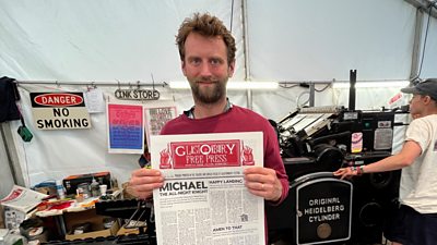 Aidan Meighan holds the Glastonbury Free Press in front of the Heidelberg press it's printed on