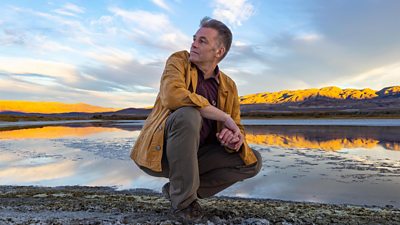 Chris Packham crouches beside a lake in a remote location. The sunrise illuminates mountains in the distance behind him.