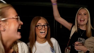 Three women celebrate England's Euros win, one with her arm in the air in celebration.