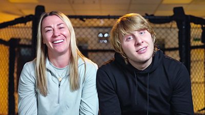 Two young people sat in front of a metal cage designed for UFC fighting