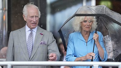 King Charles points and Queen Camilla in a light blue dress smiles while she holds an umbrella.