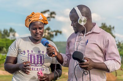 鶹ƵAV Media Action interviewing a farmer for a radio programme in East Africa - the women is wearing a yellow headscarf and the journalist is wearing a pink striped shirt. She is gesturing with her hand as she talks.