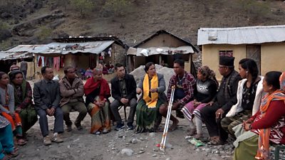 A group of people in Nepal are sitting together outside of their houses, we can see damage and some fallen rocks on the roofs caused by an earthquake. There is a lady in a yellow scarf and one man with crutches who is living with a disability. There are seven women and five men they are listening to the lady wearing the yellow scarf.