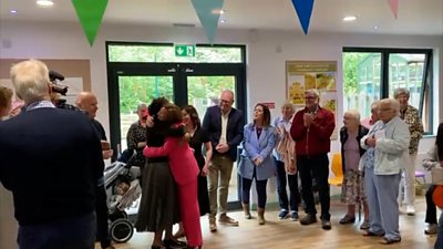 Group of people standing in a community hall, in the middle is Eluned Morgan wearing a pink suit and hugging another woman