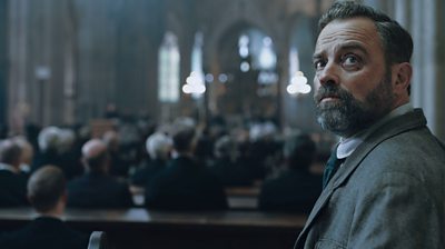 Juergen Maurer (Oskar Rheinhardt) standing at the back of a church looking up at something off screen. Blurred people in seats in background