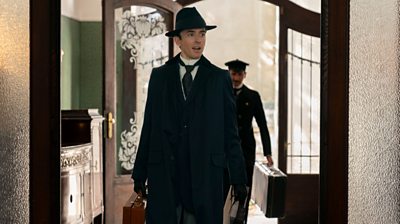 Matthew Beard (Max Liebermann) in a suit, coat and hat carrying a suitcase. He's walking through a hallway with a concierge behind him