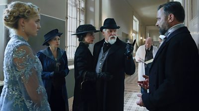 Characters in a hallway looking concerned or cross. Conleth Hill (Mendel Liebermann) is pointing his finger at a man