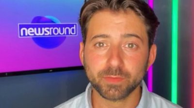 Ricky Boleto, with a short-cropped beard and wearing a grey polo shirt, sits in front of a pink and blue Newsround logo