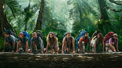 The Celebrity Race Across The World contestants pose ready to run at the starting line, crouched on the ground wearing backpacks in a rainforest setting