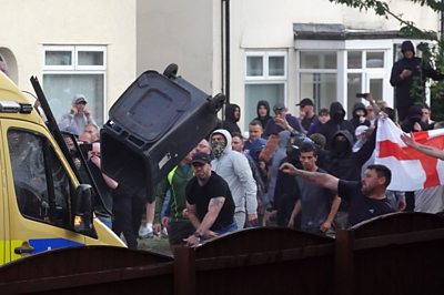 Men, some wearing masks or holding an England flag, throw a wheelie bin at a police van