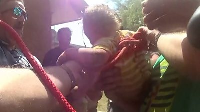 There are men surrounding a toddler who has rope wrapped around him after being rescued