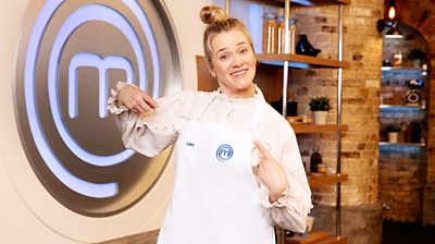 Edith Bowman in the MasterChef kitchen wearing an apron in front of the MasterChef logo