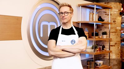 Ian ‘H’ Watkins in the MasterChef kitchen wearing an apron in front of the MasterChef logo