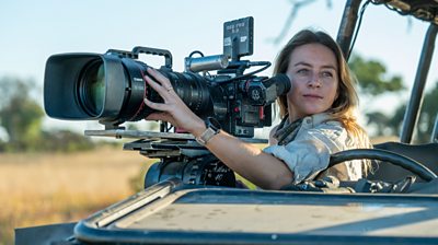 Woman in the front of a car with a huge camera looking at something off screen