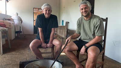 Scott Mills and his husband Sam sit on wooden chairs wearing plastic caps, stirring something in a large metal bowl