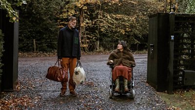 Abe (Darren Boyd) and Freya (Kyla Harris) look to each other in the driveway of a house. Abe is holding two large holdalls, while Freya wears a large fluffy coat. Autumnal leaves litter the ground. A large entrance gate with a keypad marks the entry to the driveway. 