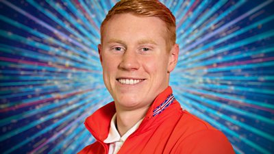 Tom Dean wears a red jacket with a Union Jack on the collar over a white shirt. He stands against a blue glittering background.