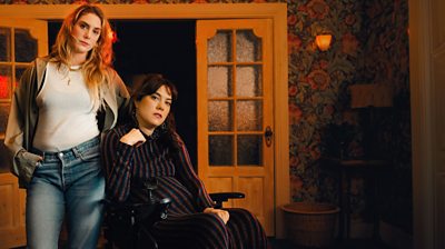 Key Art. Characters Jo (Elena Saurel) and Freya (Kyla Harris) look to camera from a living room. 