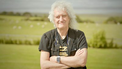 Brian May standing in a field with his arms crossed