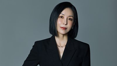 Hee Jun Yoon wears a black suit and stands against a grey backdrop