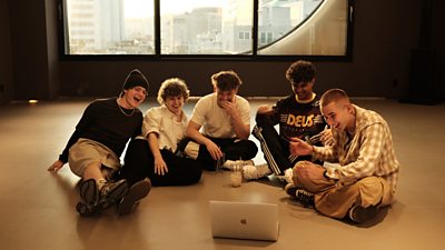 The band sit on the floor in rehearsals laughing at a laptop screen