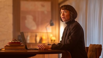 Anna Maxwell Martin at a table with a laptop in front of her. She's looking slightly up with a pensive expression