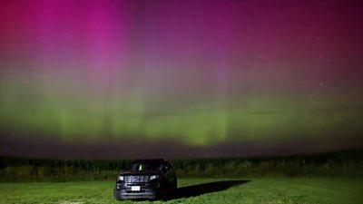 Pink and green Northern Lights hang in the sky above a car in a field at night