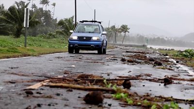 A blue car drives drives over a road littered with branches next to the sea