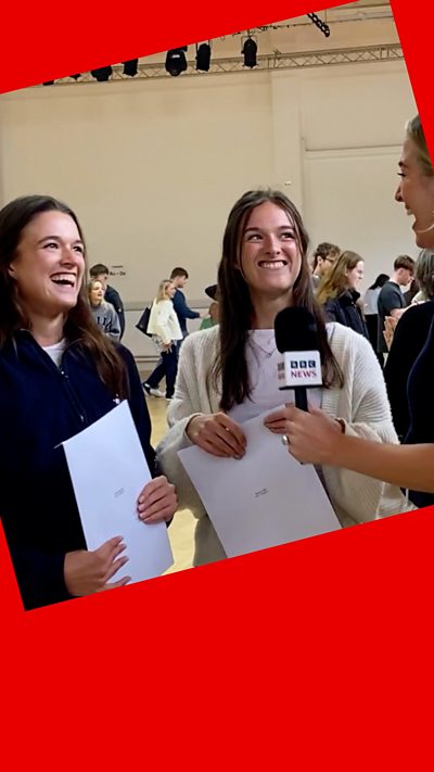 Twins smile - they have long dark hair and are holding their results papers