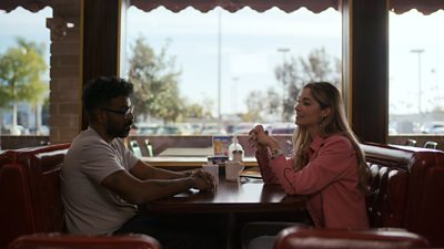 Romesh Ranganathan and Dr Julia Shaw sitting opposite sides of a table in a diner