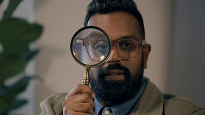 Romesh Ranganathan looking to the camera with a magnifying glass to his face