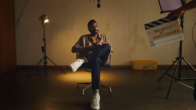 Behind the scenes shot of Romesh Ranganathan sitting on a chair with lighting behind him and a clapperboard in the foreground