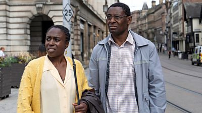 Sharlene Whye and David Harewood as Pam and Denis Bottomley looking off to the left at something offscreen on a high street 