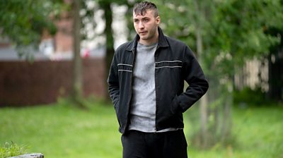 Oliver Huntington as Ryan Bottomley with his hands in his jacket pocket and wet hair. Grass and tree in background