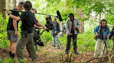 behind the scenes shot of David Harewood and Bethany Asher in a forest with lots of film and sound crew