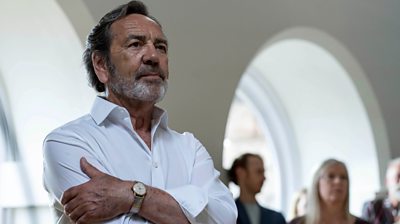 Robert Lindsay as Franklin Warner with his arms crossed looking at something offscreen