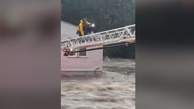 A woman and man crawl along a fire engine ladder above rushing water