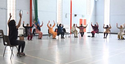 10 women sit on chairs in a sports hall with their arms raised