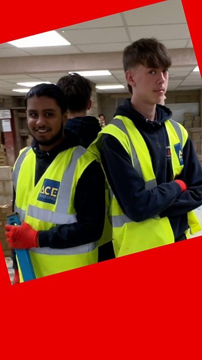 Two trainee bricklayers, wearing high-vis jackets, look over their shoulder and smile at the camera.