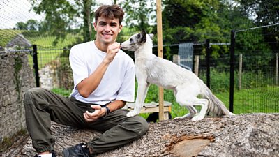 a man sitting on a tree trunk feeding a white fox and smiling to camera