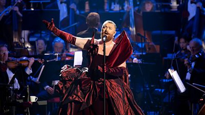 Sam Smith wearing an elaborate burgundy velvet gown singing with an orchestra in the background