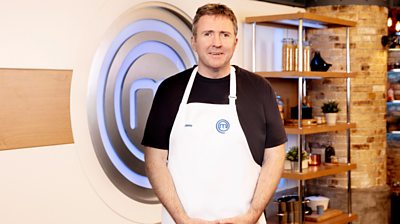 Jamie MacDonald in the MasterChef kitchen wearing an apron in front of the MasterChef logo