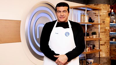 Tamer Hassan in the MasterChef kitchen wearing an apron in front of the MasterChef logo