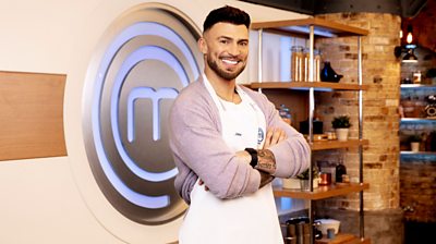 Jake Quickenden in the MasterChef kitchen wearing an apron in front of the MasterChef logo