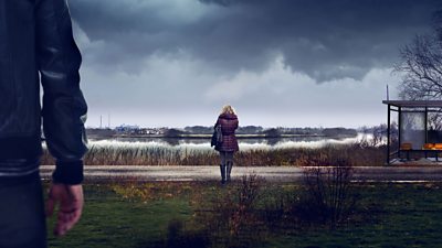 composite image showing the back of a woman in a winter coat in the distance looking out to a lake. man's arm and side in the foreground implied he's looking at her. weather gloomy and grey